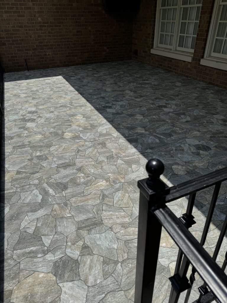 A sunlit patio with a natural stone tile floor and a black metal railing in the foreground.